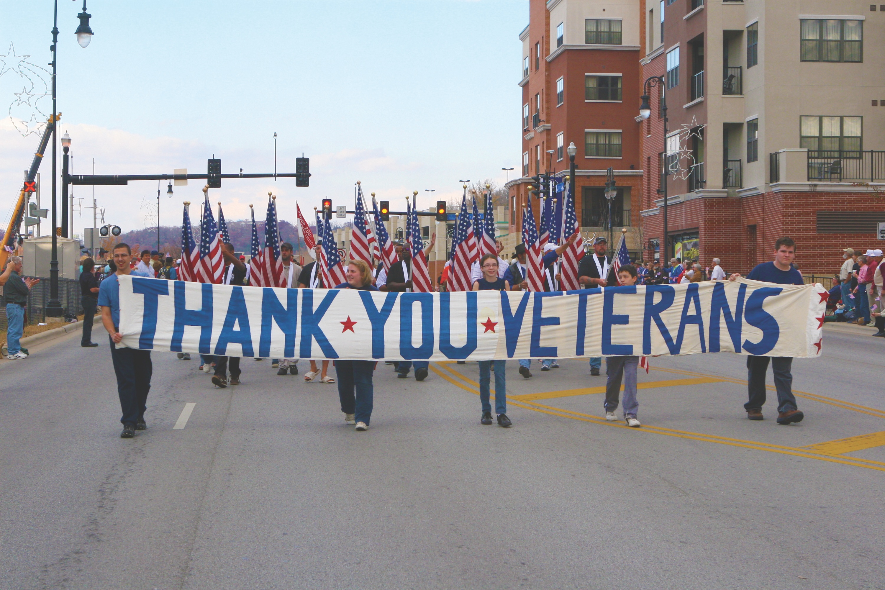 Parade participants carrying American flags and a large 'Thank You Veterans' banner down a city street during a Veterans Day celebration in Branson, MO
